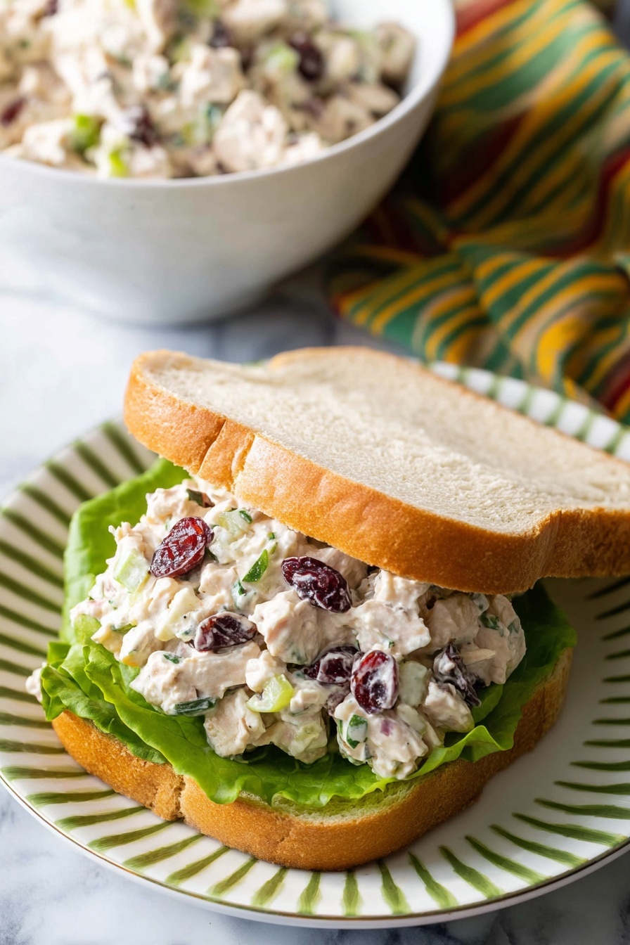 A sandwich with two slices of light brown bread, one slice on top slightly tilted, placed on a white plate with green stripes, on a white marbled surface. Inside the sandwich, there is a fresh green lettuce leaf at the bottom, topped with a chunky mixture of creamy white chicken salad with visible small pieces of celery, dark red dried cranberries, and flecks of green herbs. In the background, there is a white bowl filled with more of the chicken salad, and a striped cloth with green, yellow, and red colors. Photo taken with an iphone --ar 2:3 --v 7