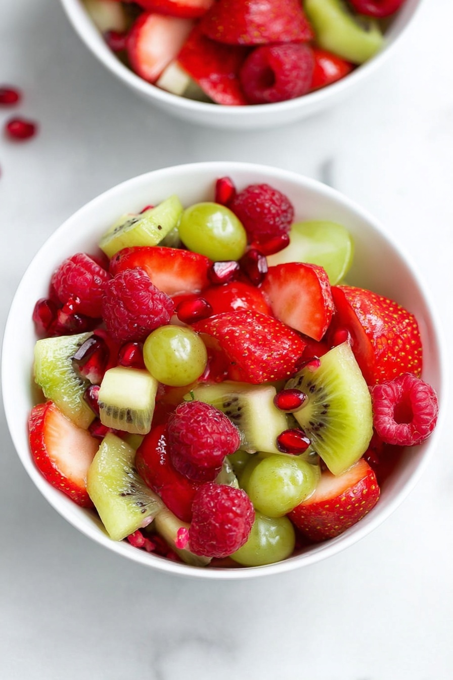 A white bowl filled with a colorful fruit salad shows a mix of layers: bright red strawberries sliced in halves, red raspberries with their bumpy texture, small green grapes cut in halves, and green kiwi pieces with tiny black seeds visible. There are small scattered red pomegranate seeds that add pops of color. The bowl is set on a white marbled surface with another similar bowl partially visible in the background. Photo taken with an iphone --ar 2:3 --v 7