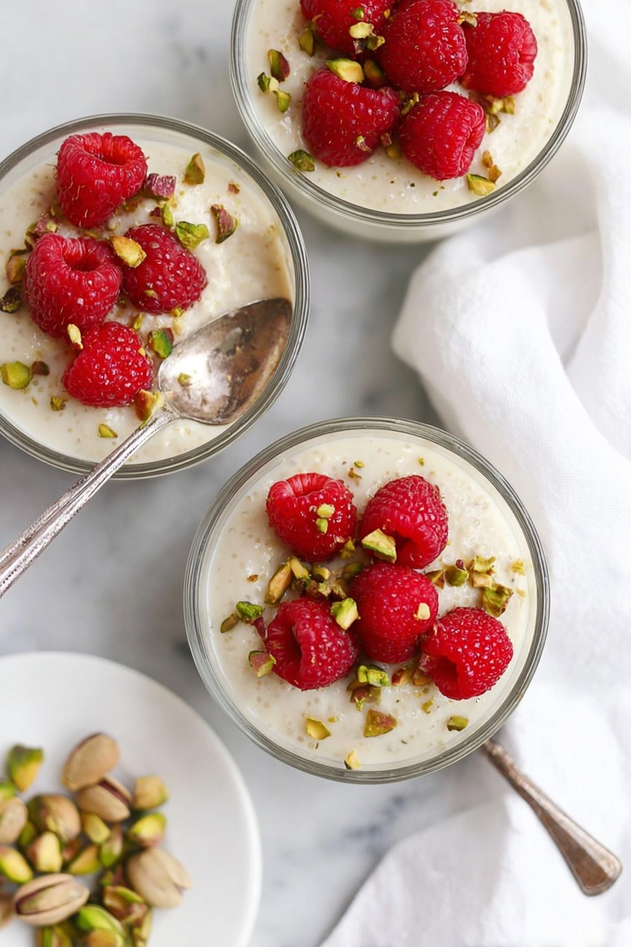 Three small glass bowls each hold a creamy white pudding as the base layer, topped with several bright red raspberries and scattered greenish-brown chopped pistachios. One bowl in the center has a silver spoon resting inside. The bowls sit on a white marbled surface with a white cloth nearby and a small white plate holding more pistachios in the corner. The colors are soft and fresh, with the red berries providing a bright contrast to the creamy pudding and green nuts. photo taken with an iphone --ar 2:3 --v 7