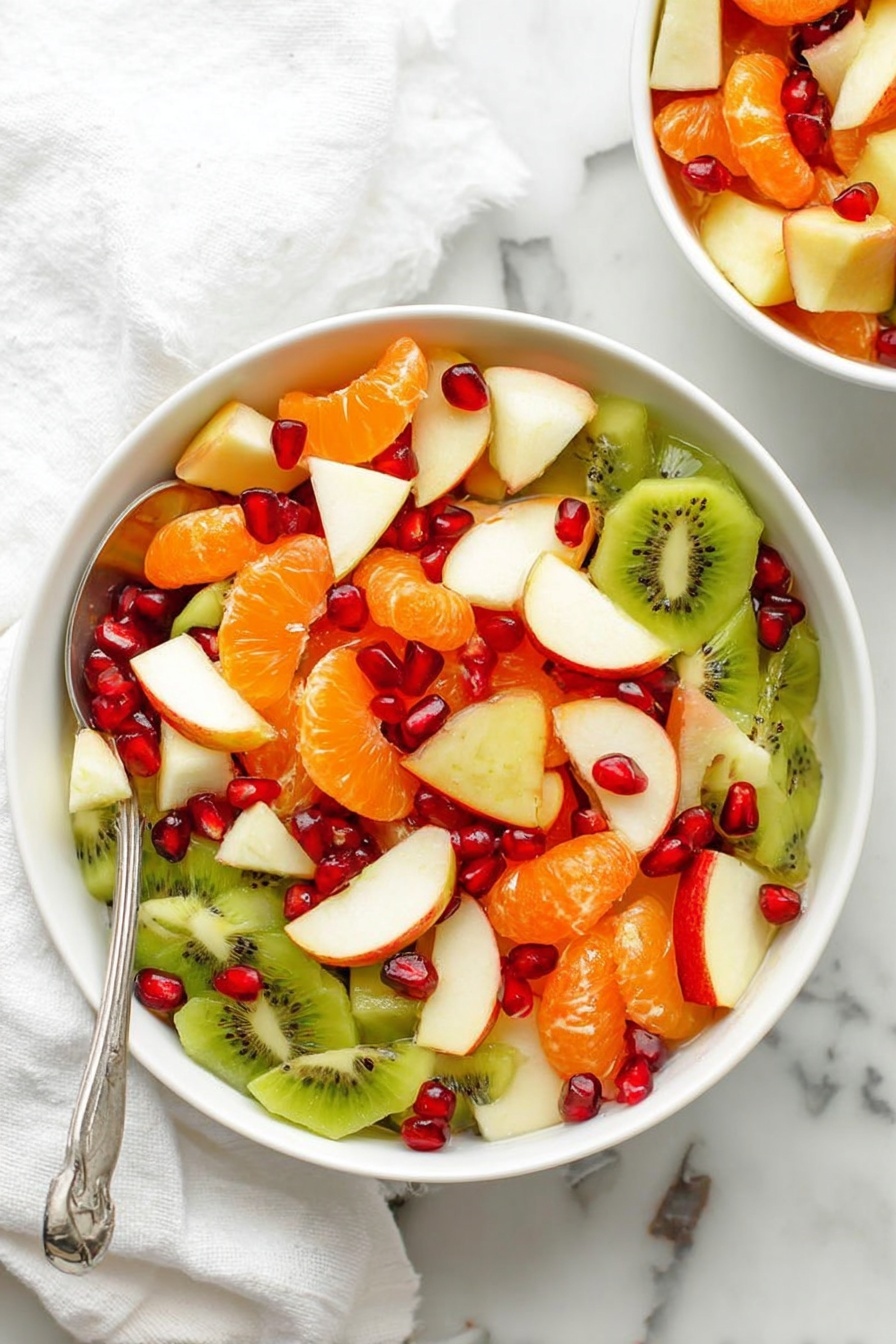 The image shows a white bowl full of colorful fruit salad placed on a white marbled surface. The salad has three main layers: the bottom layer is made of green kiwi slices with black seeds, the middle layer has segmented orange tangerines, and the top layer has white apple slices with red skins and scattered bright red pomegranate seeds. A silver spoon is inside the bowl at the bottom left. A white cloth is partly visible to the left side. Part of another white bowl with the same fruit salad is seen near the top right corner. photo taken with an iphone --ar 2:3 --v 7