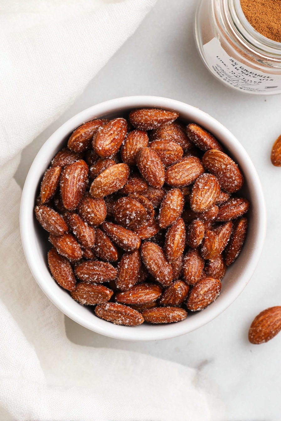 A white bowl filled with shiny, brown almonds covered in a light layer of sugar and cinnamon powder. The almonds are whole and rough-textured, filling the bowl completely and looking crunchy. The bowl is placed on a white marbled surface with soft white cloth partially visible around it, and a small glass jar of cinnamon powder with a simple label is placed near the top right corner. The overall look is clean and simple with a focus on the warm, brown nuts. photo taken with an iphone --ar 2:3 --v 7