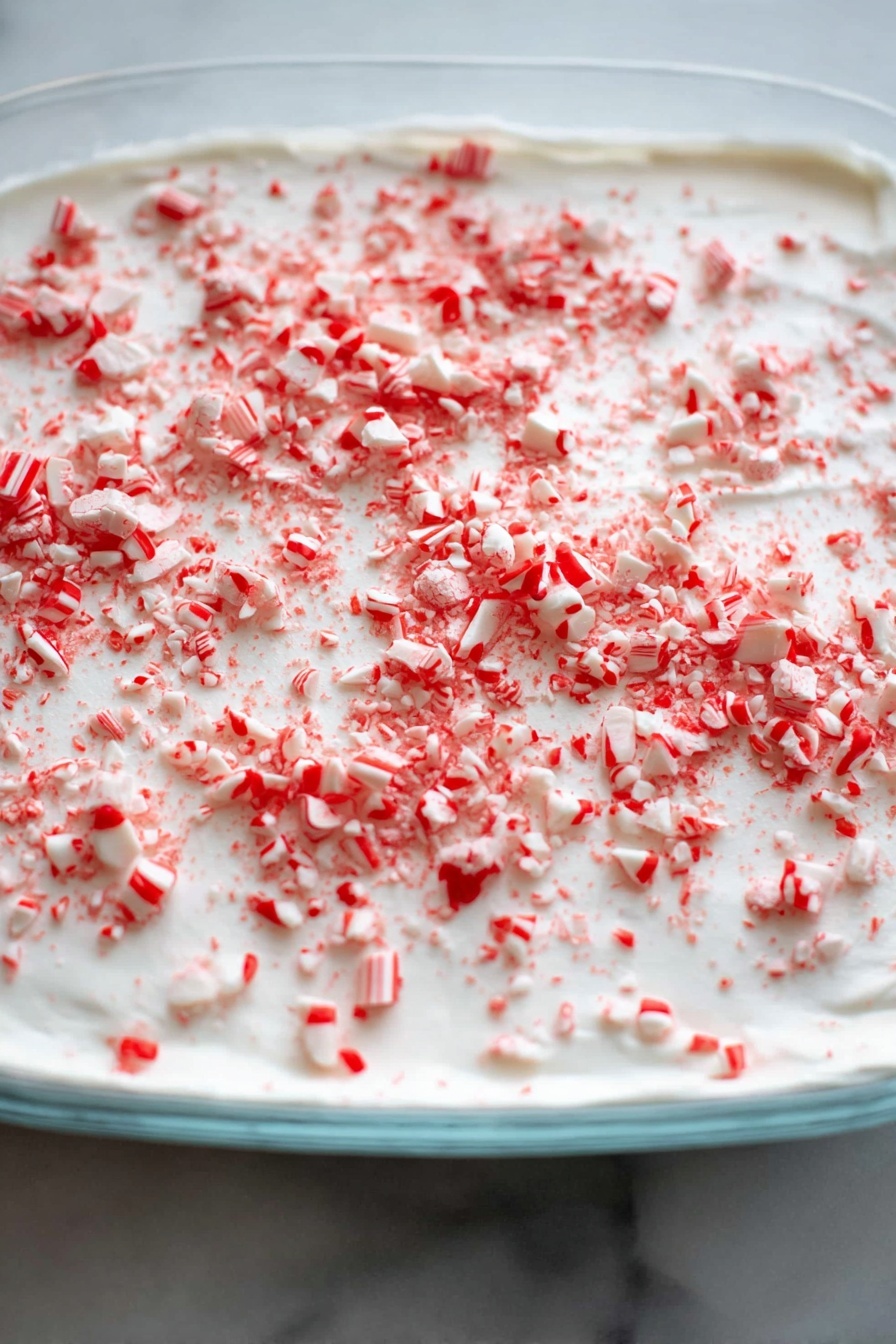 The image shows one thin layer of white cream spread evenly in a shallow clear dish, topped with crushed red and white peppermint candy pieces scattered all over the surface. The candies vary in size, with some small bits and some larger chunks, adding texture and color contrast to the smooth white cream. The dish is placed on a white marbled surface. photo taken with an iphone --ar 2:3 --v 7