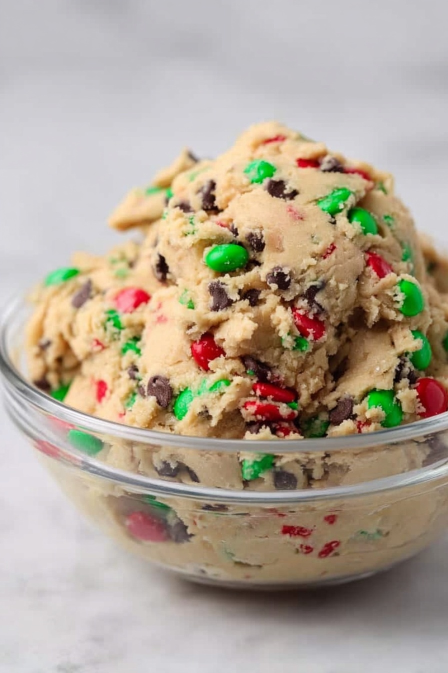 The image shows a close-up view of soft cookies stacked in a pile on a white marbled surface. Each cookie has a light beige dough base densely dotted with small dark chocolate chips and larger red and green candy-coated chocolates spread evenly across the top. The cookies have rounded, slightly uneven edges and a soft texture with tiny cracks on the surface. The candies are bright and glossy, creating a colorful contrast and adding a festive look to the warm-toned dough. photo taken with an iphone --ar 2:3 --v 7