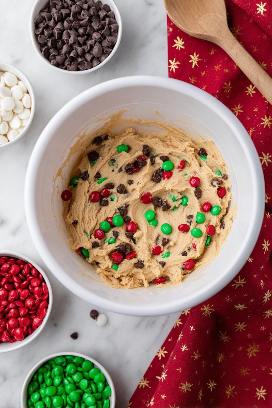 A close-up of a square cookie held by a woman's hand with black nail polish, showing a cracked surface with red, green, and white candy-coated chocolates and small dark chocolate chips embedded throughout. The cookie has a golden-brown color with a slightly rough texture and visible cracks on top. In the blurred background, other similar cookies are placed on a white marbled surface. photo taken with an iphone --ar 2:3 --v 7