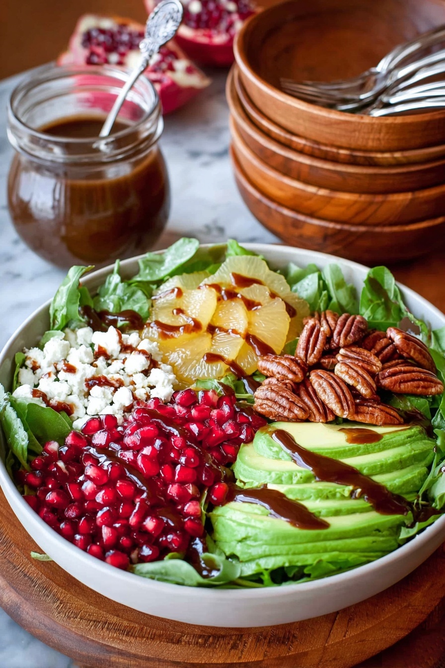 A white bowl filled with mixed green leafy vegetables forms the base layer, topped with sections of bright red pomegranate seeds, sliced yellow grapefruit segments, creamy white crumbled cheese, sliced green avocado fanned out, and a pile of dark brown pecans. A dark brown dressing is drizzled in thin lines over the cheese and avocado. The bowl sits on a wooden table next to a small glass jar holding more dressing with a spoon inside, and a stack of similar wooden bowls with metal forks resting inside them. The setting is on a white marbled textured surface photo taken with an iphone --ar 2:3 --v 7