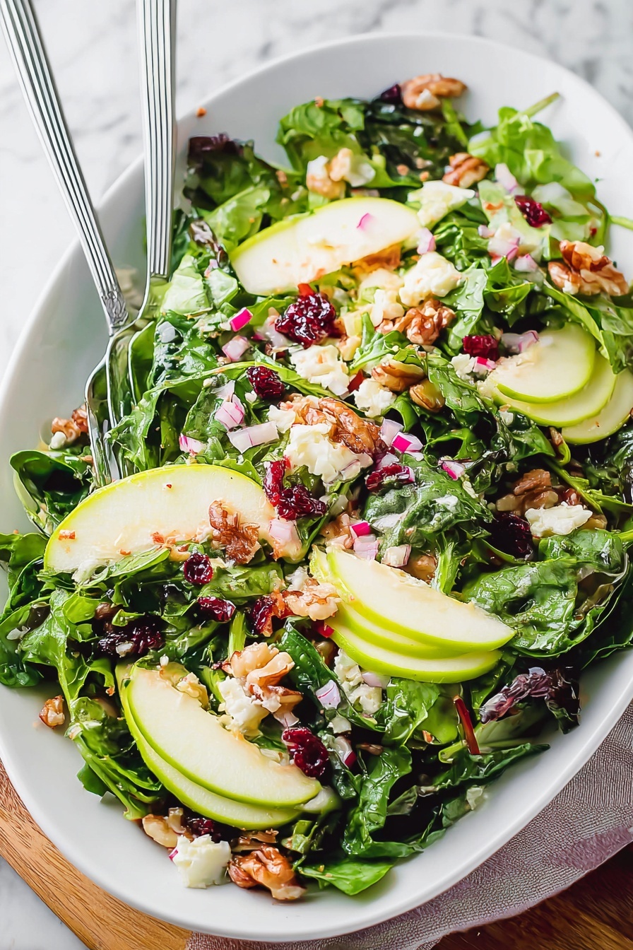 This image shows a fresh salad served in a white oval bowl placed on a white marbled surface. The salad is built in many layers, starting with a mix of green leafy vegetables including spinach and darker leafy greens as the base, giving a deep green and rich texture. On top of the greens, thin slices of light green apple are spread evenly, adding a smooth, crisp layer. Scattered around are small chunks of creamy white cheese, finely chopped pink onions, and dark red dried cranberries. Walnut pieces add a rough texture with a warm brown color scattered on top. The salad looks fresh and colorful with a light dressing that gives a slight gloss. Two metal forks rest on the salad's left side, partially buried in the leaves. photo taken with an iphone --ar 2:3 --v 7